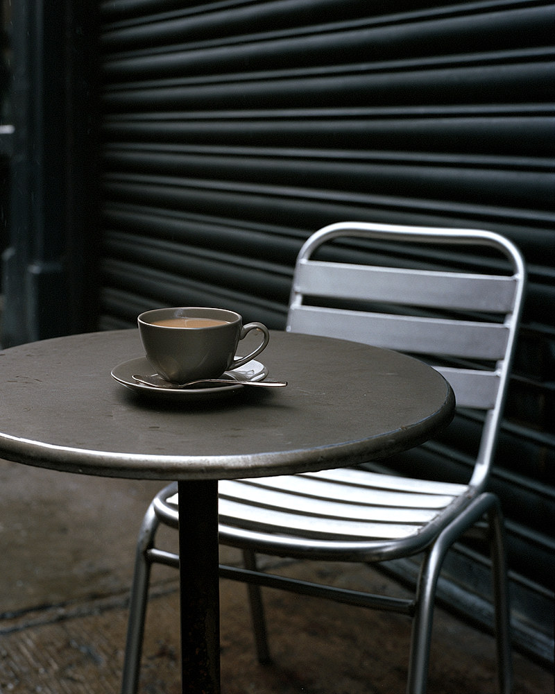 An empty caf&eacute; table with a cold cup of tea, a chair, and a metal shutter behind